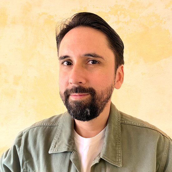 Homem branco, cabelo liso preto com barba e bigode. Ele usa uma jaqueta verde e uma camisa branca. Está sorrindo levemente.